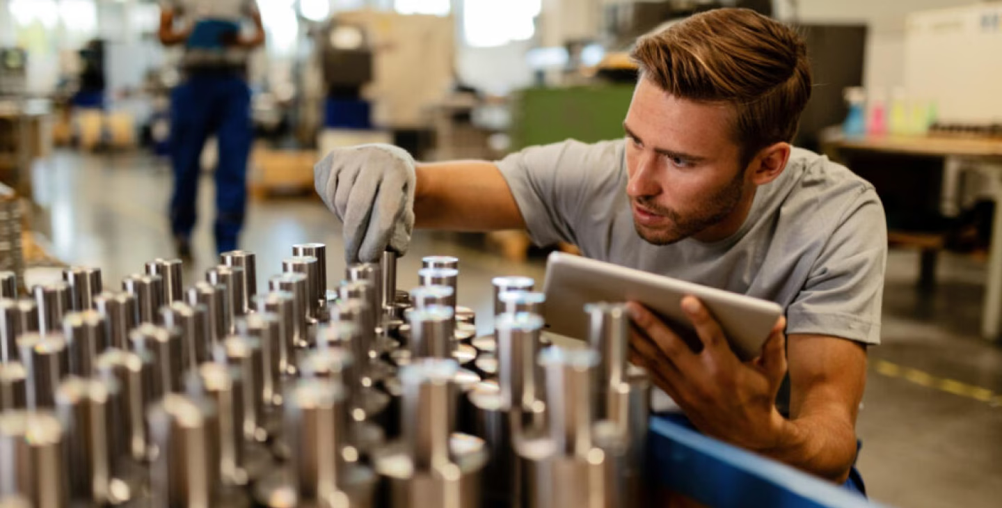 Ouvrier effectuant un contrôle qualité sur des pièces métalliques dans une usine, utilisant une tablette numérique pour vérifier les données.