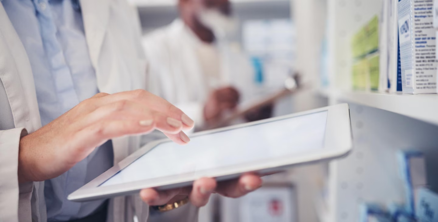 Une personne en blouse blanche utilise une tablette tactile dans un laboratoire médical, avec d'autres personnes en arrière-plan.