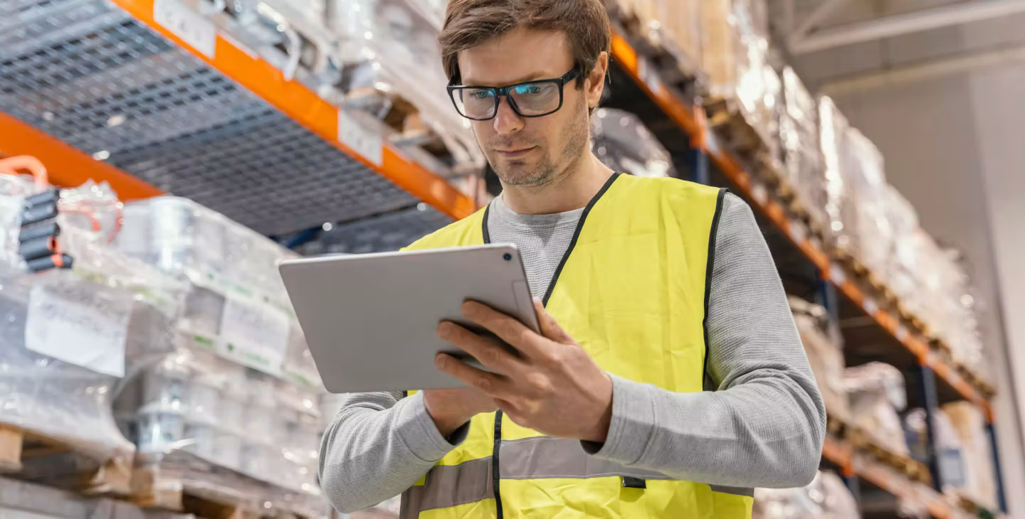 Employé en gilet jaune dans un entrepôt utilisant une tablette pour gérer l’inventaire des stocks sur les étagères.