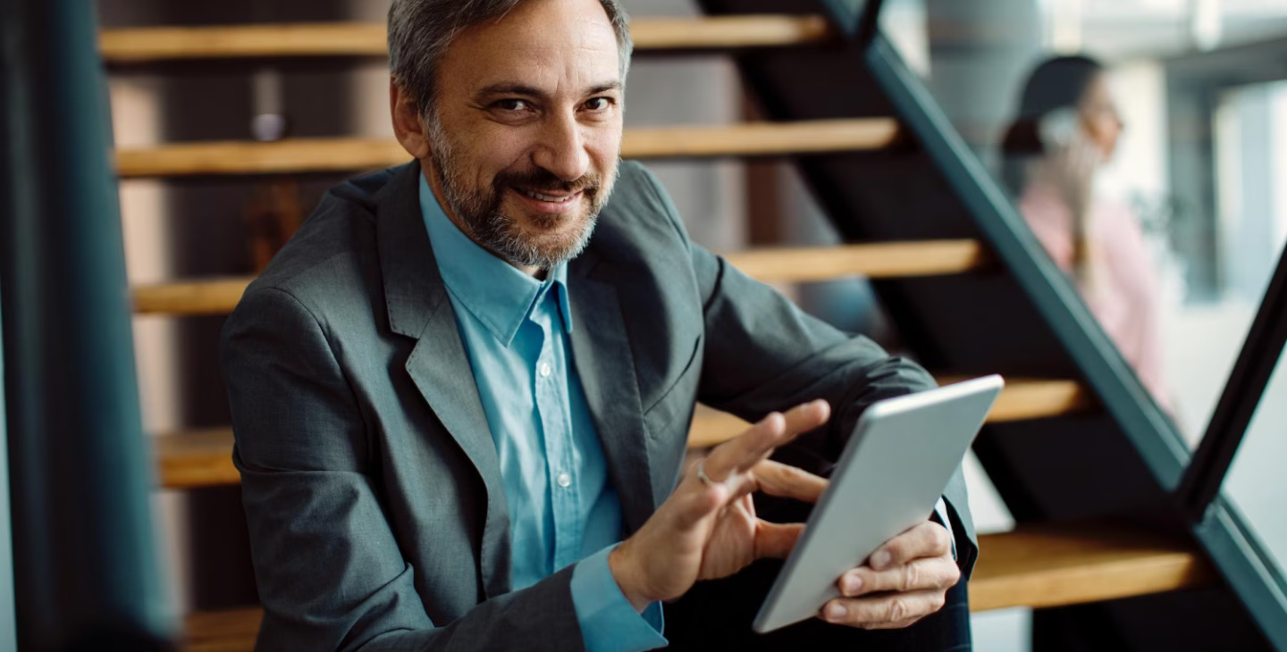 Homme souriant en costume utilisant une tablette assis sur des escaliers dans un bureau moderne.