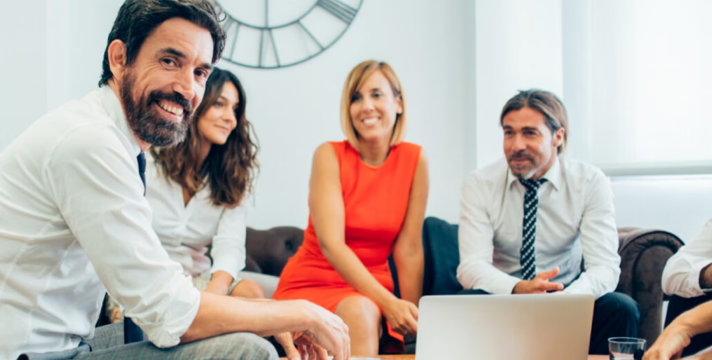 Groupe de professionnels souriants en réunion informelle au bureau, discutant autour d’un ordinateur portable dans une ambiance collaborative et détendue.