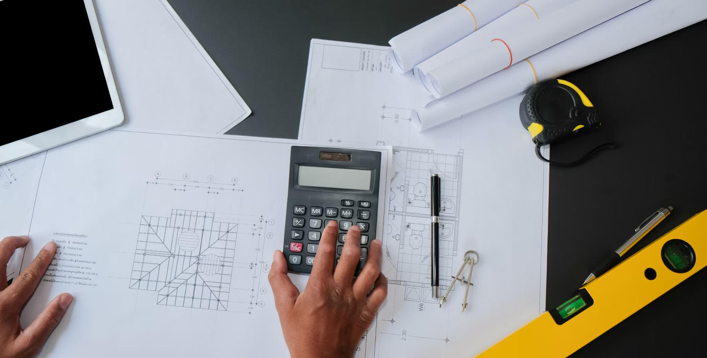 Vue de dessus d’un bureau d’ingénierie avec plans techniques, calculatrice, tablette et outils de mesure.